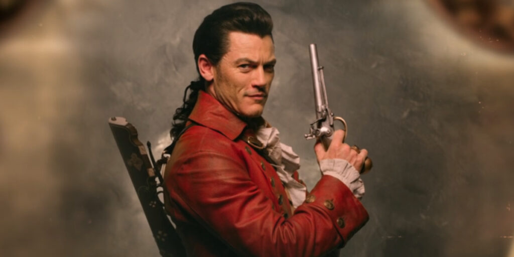 A cocky Luke Evans sits posing with a pistol in Beauty and the Beast (2017).