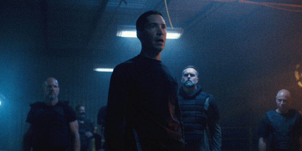 Justin Long stands shocked in a dark room surrounded by fellow cops in Night Patrol.