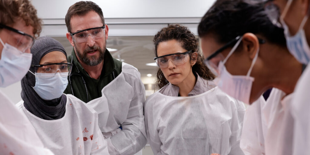 Gerran Howell, Amielynn Abellera, Noah Wyle, Sepideh Moafi, Supriya Ganesh shown huddled together in scrubs in The Pitt S2.