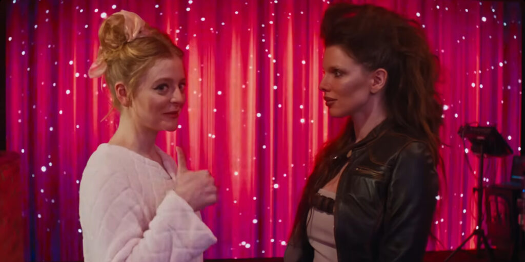 Anna Baryshnikov flashes an uneasy thumbs up at the camera while talking with Julia Fox in Idiotka.